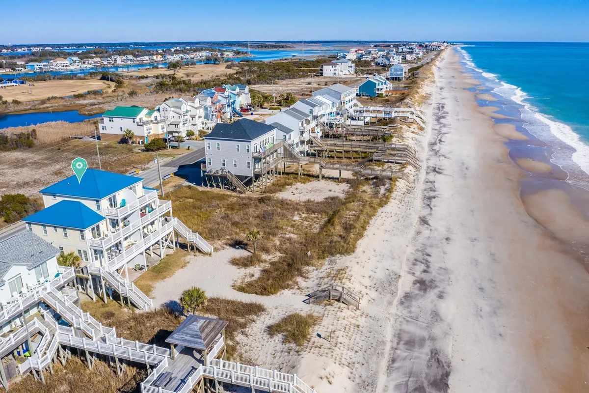 A wide exterior shot featuring beachfront houses nestled behind sand dunes, with the sparkling ocean waves and walking paths inviting guests toward the shoreline. The sky is clear and bright, evoking the lively activity of summer and the peacefulness of off-season mornings.