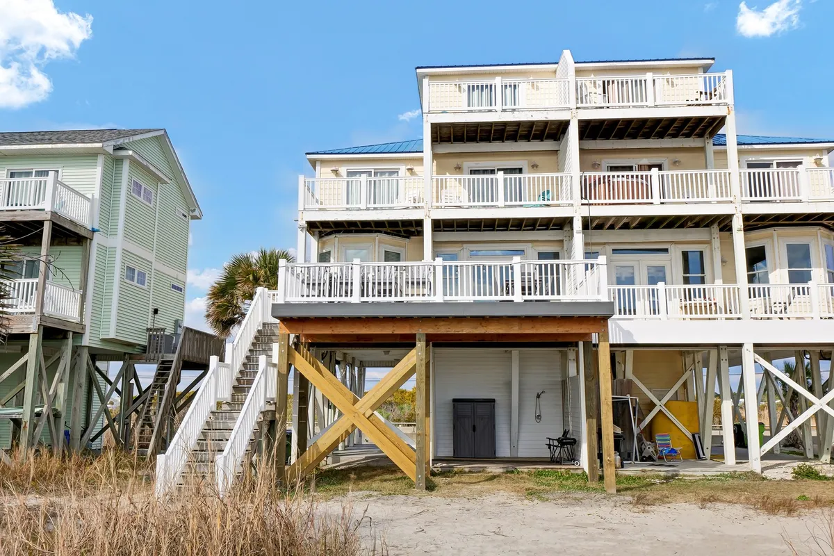 An exterior view of a multi-level beach house with a visible outdoor staircase, palm trees, and an outdoor seating area. The image highlights access points, parking (potentially where the EV charger is located), and the home's overall accessibility.