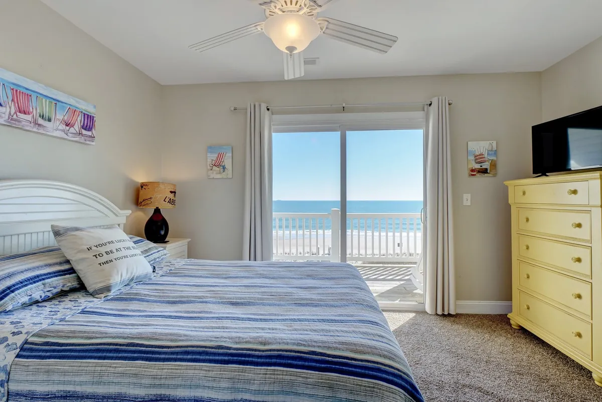 This image features a cozy bedroom with a bed dressed in striped bedding, a large window offering a beautiful ocean view, a yellow dresser, and a ceiling fan. Natural light floods the room, creating a calm and restful ambiance—perfect for unwinding after a day in the sun.