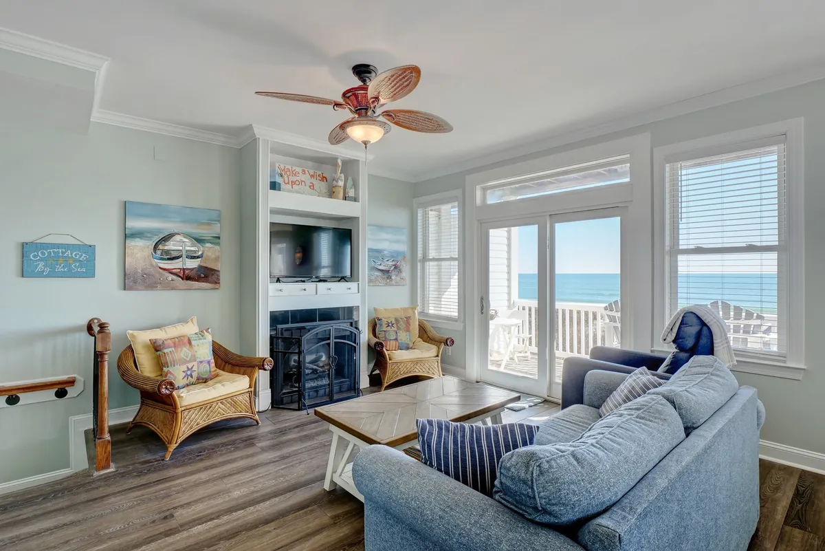 This cheerful living room features a cozy blue sofa, colorful accent chairs, and large windows overlooking the ocean. The space is arranged for group activities like board games or enjoying scenic sunsets, reflecting a family-friendly atmosphere with plenty of natural light.