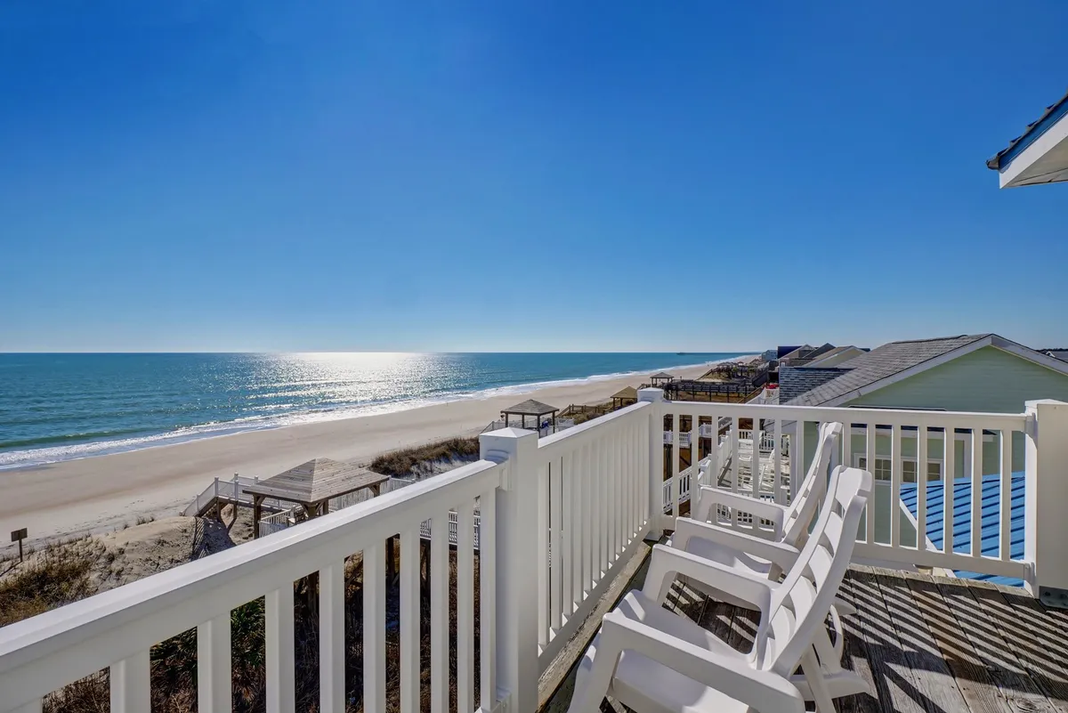 A spacious wooden deck overlooking the beach and ocean, complete with deck chairs positioned for both relaxation and dining. The sand and water are visible in the background, creating an airy and open connection to the coastal surroundings.