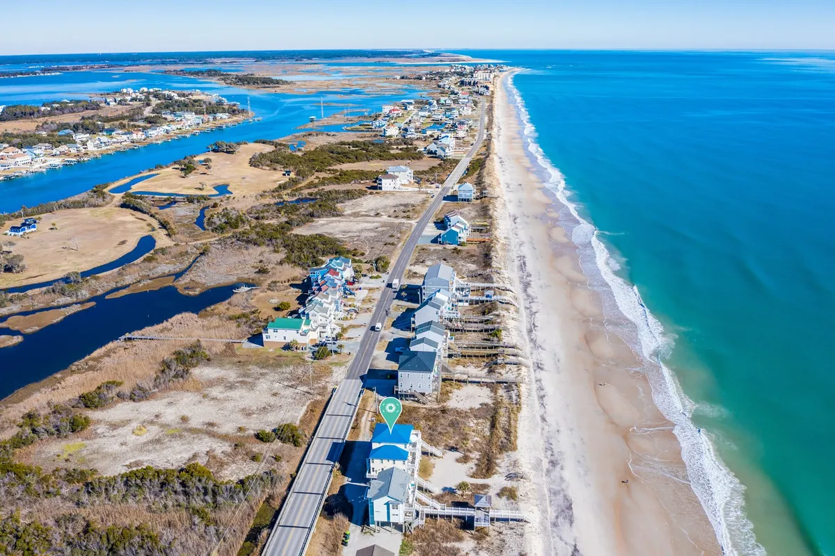 A sweeping view of North Topsail’s beachfront houses lining the coast, a coastal road, blue ocean stretching out towards the horizon, and scenic waterways winding through the landscape. People can be seen enjoying relaxed walks and beachcombing along the wide shore.