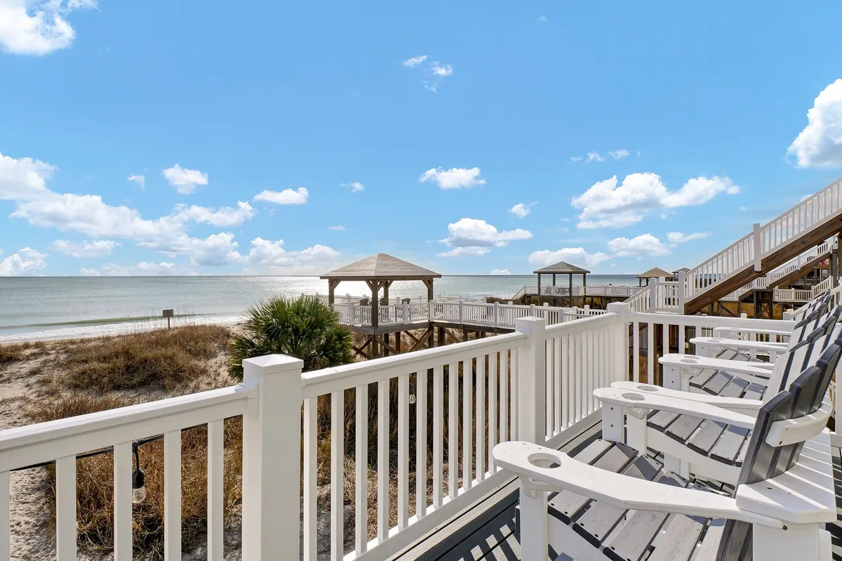 A tranquil beachfront deck with white railings and classic Adirondack chairs overlooking the ocean, with a gazebo visible in the background. The setting is sunlit and peaceful, ideal for relaxing after an outdoor adventure.