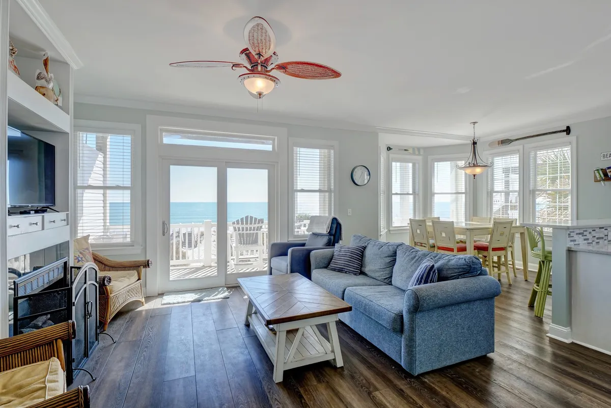 A spacious, sun-filled living area with comfortable seating, a dining area, and large windows that offer sweeping views of the Atlantic Ocean. The room exudes a casual and inviting atmosphere, perfect for gatherings and relaxation. The layout encourages connection and features a beach motif ceiling fan, underscoring the seaside vibe.