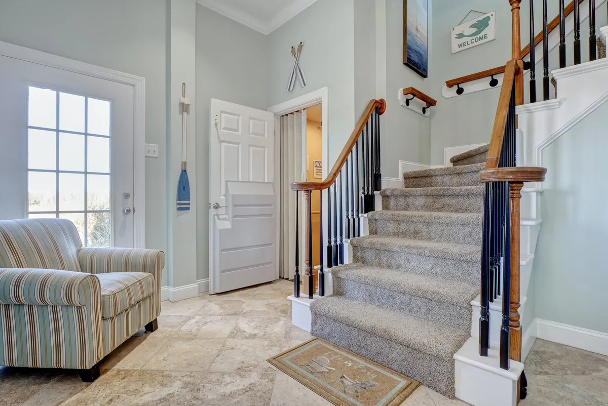 A welcoming interior view with a staircase, a striped chair, a 'welcome' sign, and decorative oars. The space reflects the entry and circulation areas of the home, possibly connecting different floors, which relates to the elevator feature.