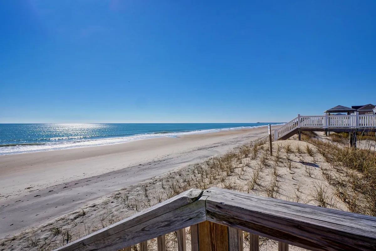 A serene exterior view of the beach and ocean with a boardwalk cutting through dune grass, leading towards the water. The scene radiates a quiet, natural beauty, perfect for shelling, wildlife watching, and enjoying the refreshing ocean breeze.