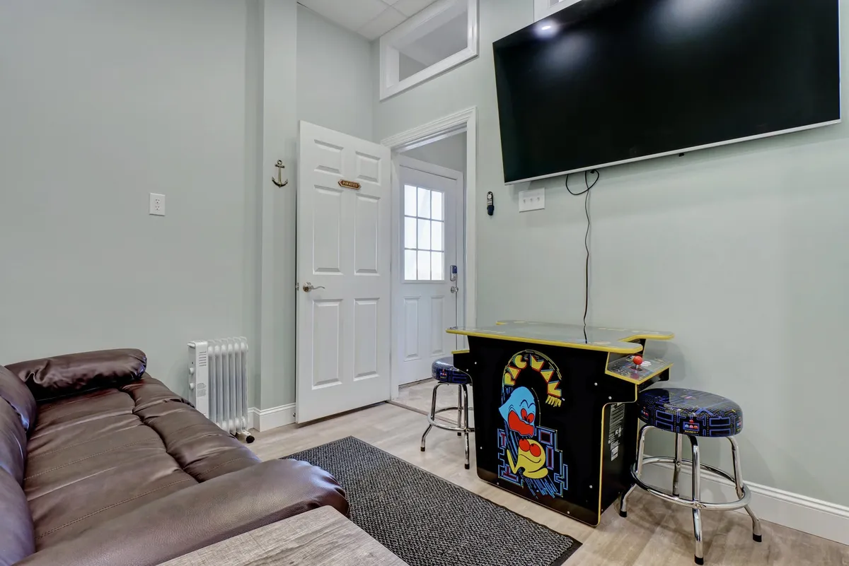 A casual game room interior featuring a brown leather couch, an arcade-style gaming table, and a large flat-screen TV. The space is designed for leisure and entertainment, with comfortable seating and an accessible layout. The door to the outside hints at easy transitions between relaxation and adventure.