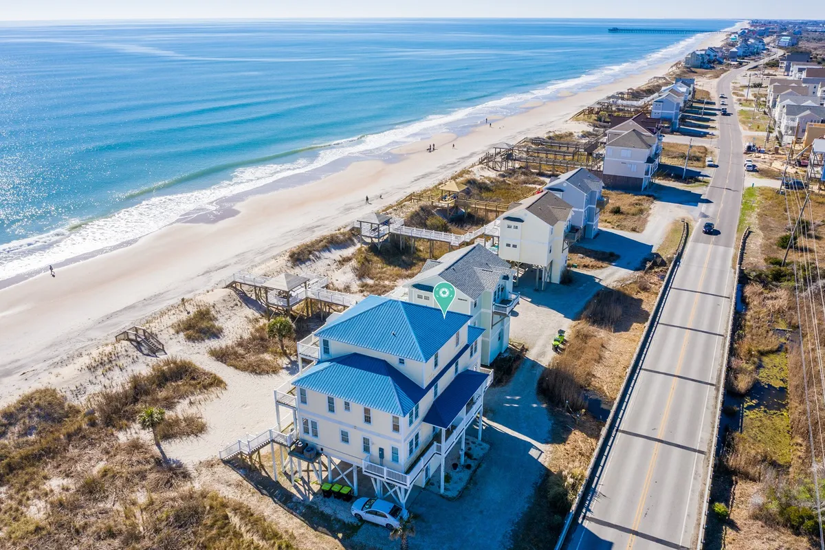 A wide exterior view of the beachfront property, showcasing its prime location right on the white sandy beach with the blue ocean and a coastal road in the background. The image gives context to the property's oceanfront positioning and ease of direct beach access.