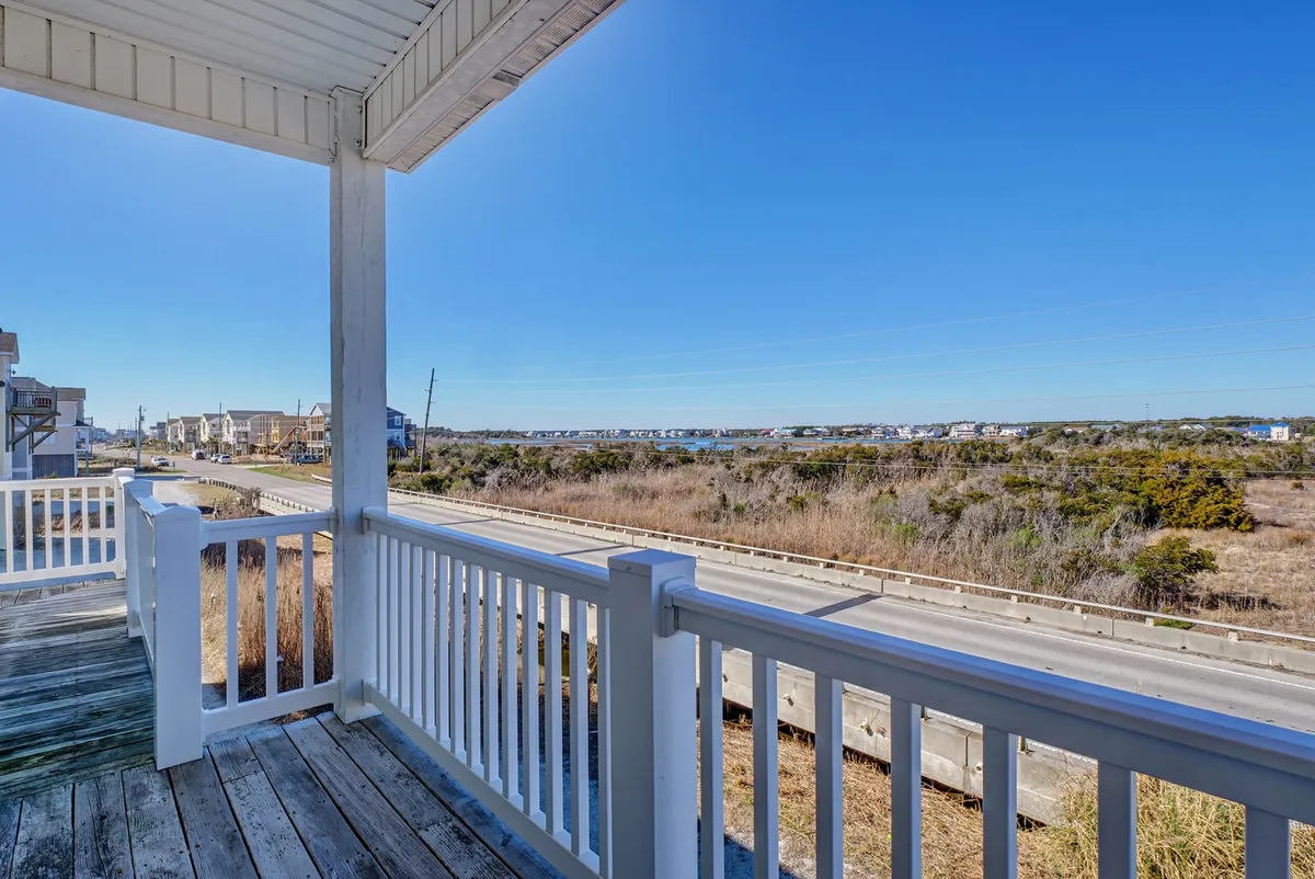 A peaceful balcony scene with a railing, road views, lush vegetation, and neighboring beach houses in the distance. The seating is ideal for enjoying morning coffee or casual meals while absorbing the coastal neighborhood ambiance.