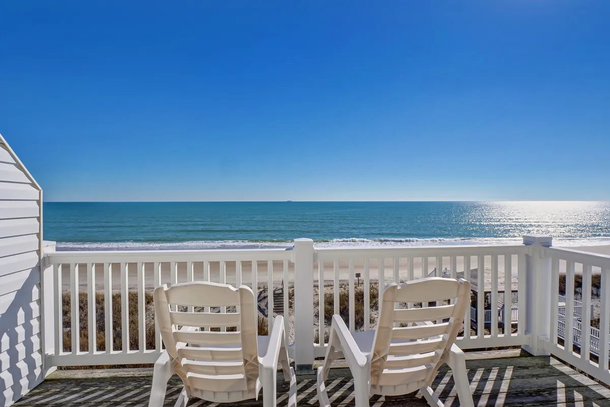 The image showcases a serene beachfront setting with two inviting white chairs positioned on an oceanfront deck, overlooking the sparkling Atlantic with a clear blue sky above. The scene exudes tranquility and showcases direct access to sand and sea.