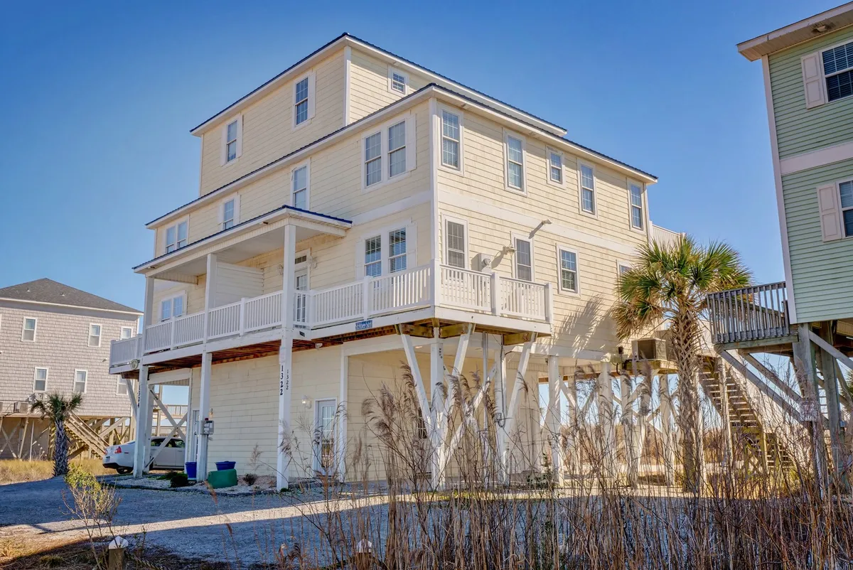 A multi-level beach house is surrounded by lush palm trees and greenery, with an expansive wooden deck set up for outdoor dining and relaxation. The setting captures guests enjoying time together, whether hosting a barbecue, laughing under the stars, or soaking in the tranquil atmosphere.