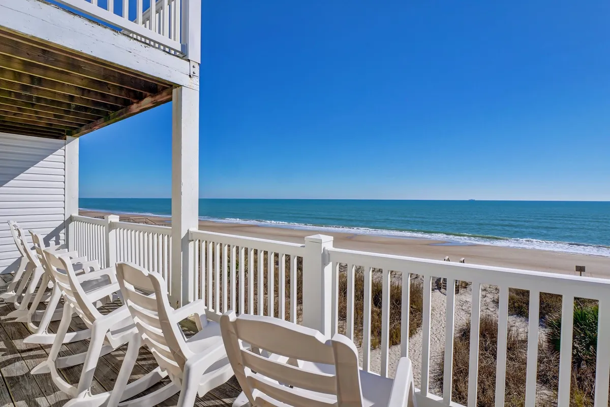 A vibrant balcony scene features seating overlooking a stunning ocean view and clear blue sky. The space is perfect for groups to socialize, enjoy a soak in the hot tub (not pictured but implied), or simply take in the seaside ambiance together.